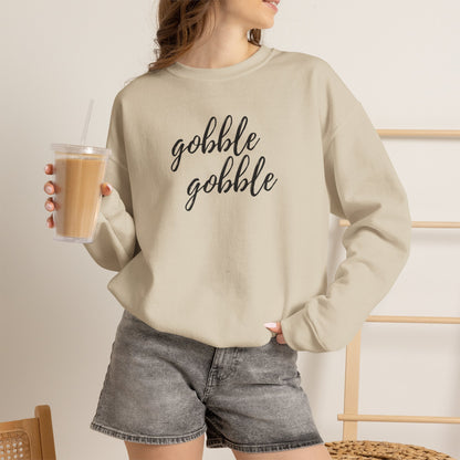 A woman wearing a beige crewneck sweatshirt with a graphic that reads 'Gobble Gobble' in black. She is holding a iced coffee and wearing gray denim shorts in a brightly lit indoor setting.