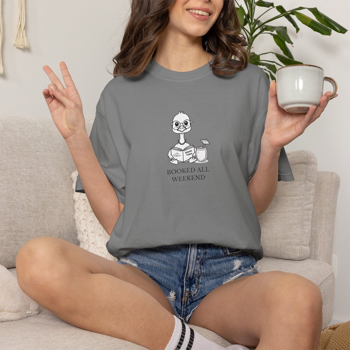 A woman sitting on a beige couch wearing a grey graphic t-shirt with a cartoon nene goose reading and the text 'BOOKED ALL WEEKEND'. She is holding a white ceramic mug and making a peace sign.
