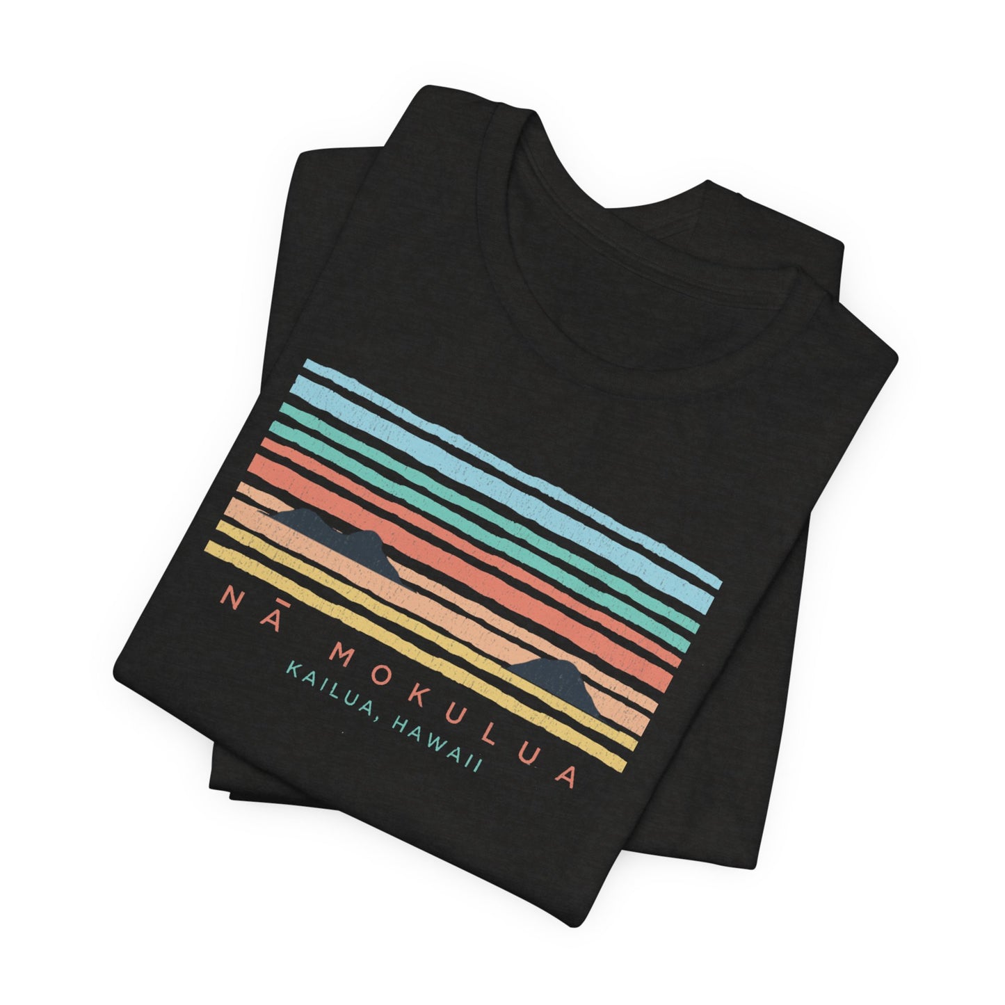 A folded black t-shirt featuring a retro sunset graphic with horizontal pastel stripes in blue, green, orange, and yellow, along with the text 'NÁ MOKULUA' and 'KAILUA, HAWAII'.