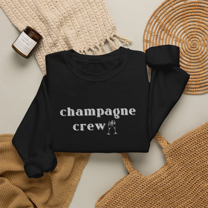 A black crewneck sweatshirt with the word 'champagne crew' printed in white lowercase serif letters across the chest, styled in a flat lay with a woven cream throw, a small amber candle jar, and a woven mat on a light beige background.