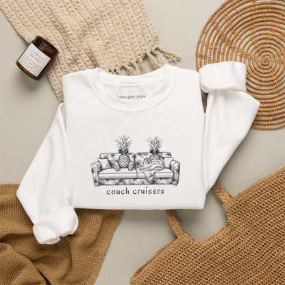 A white crewneck sweatshirt laid flat on a neutral background, featuring a black line art graphic of a couch with pineapples and the text 'couch cruisers'. The sweatshirt is styled with a beige knit blanket, a woven placemat, a straw bag, and a small candle.