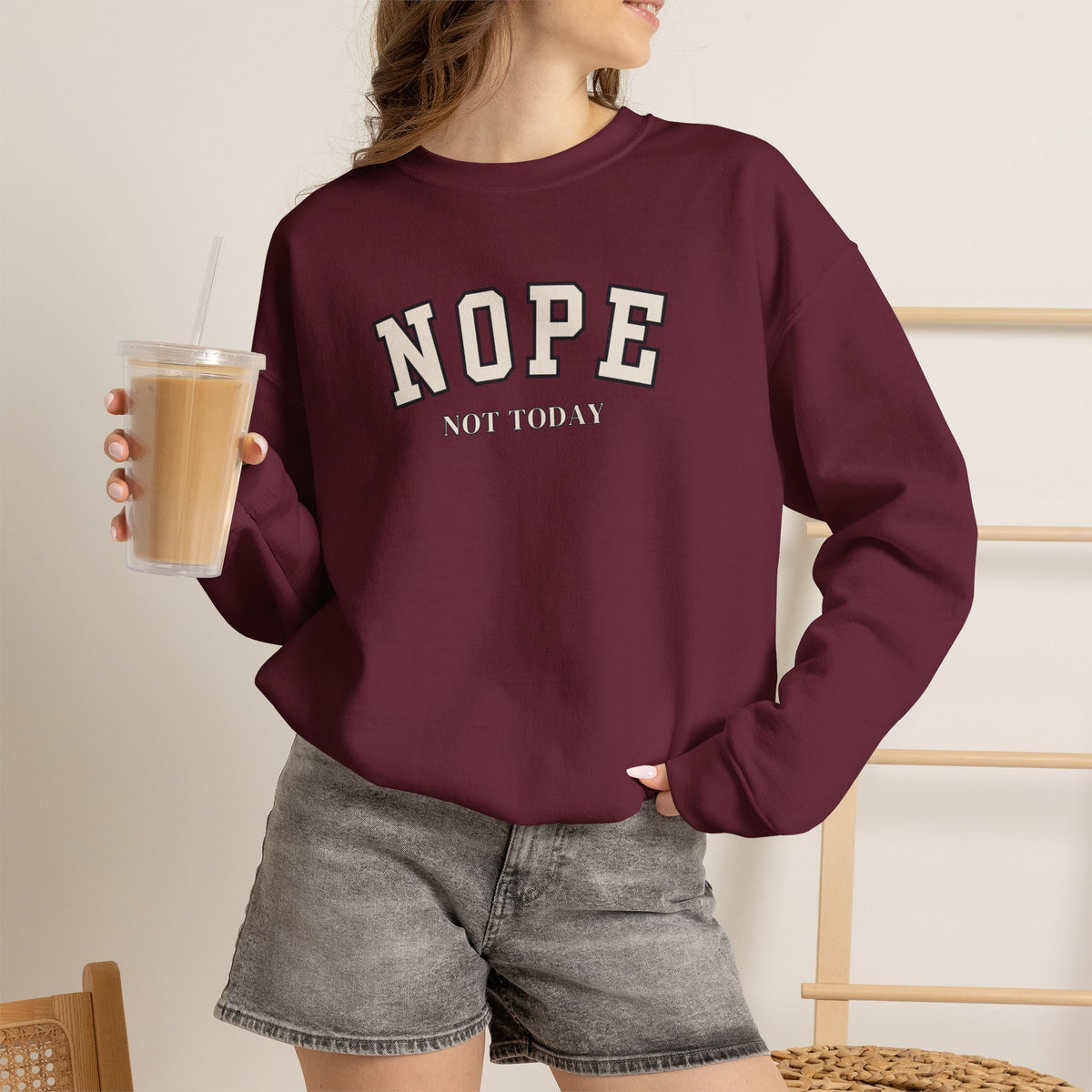 A woman wearing a maroon crewneck sweatshirt with black text reading 'NOPE NOT TODAY', paired with gray denim shorts, holding an iced coffee.