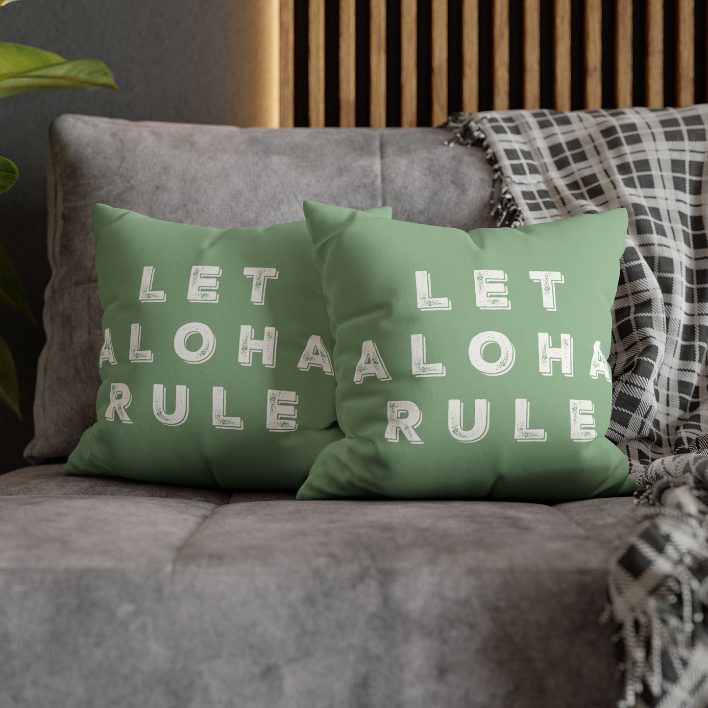 Two sage green throw pillows with white text reading 'LET ALOHA RULE' arranged on a gray sofa with a plaid throw blanket and wooden slat wall in the background.
