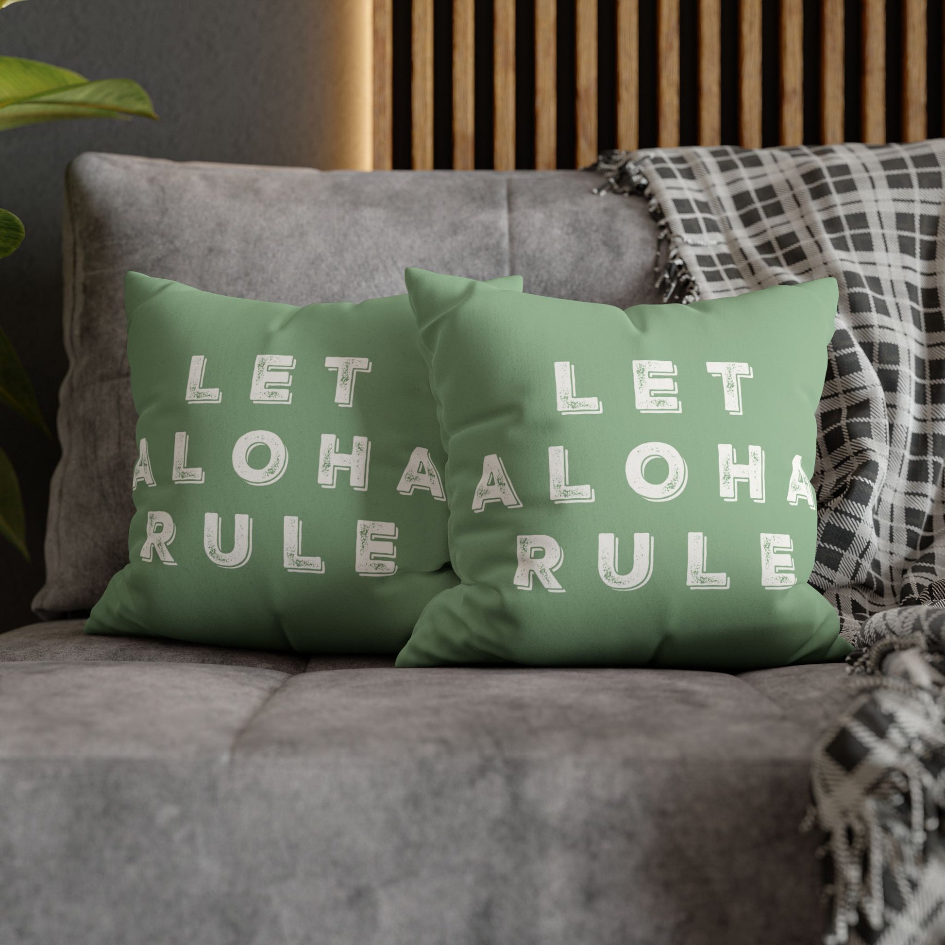Two sage green throw pillows with white text reading 'LET ALOHA RULE' arranged on a gray sofa with a plaid throw blanket and wooden slat wall in the background.