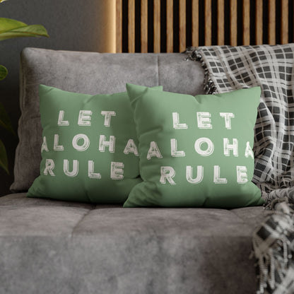 Two sage green throw pillows with white text reading 'LET ALOHA RULE' arranged on a gray sofa with a plaid throw blanket and wooden slat wall in the background.