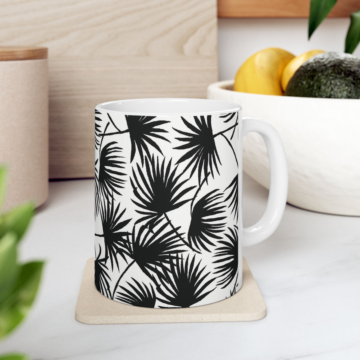 Palm Fan Black coffee mug with black and white tropical palm print by Global Village Kailua sitting on a coaster on a white kitchen counter with fruit bowl.