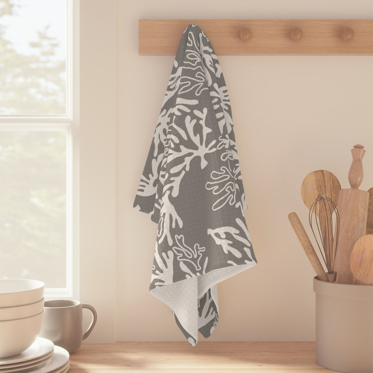 A gray kitchen towel with a white coral pattern hangs from a wooden peg on a kitchen wall, with a window, bowls, and wooden utensils in the background.
