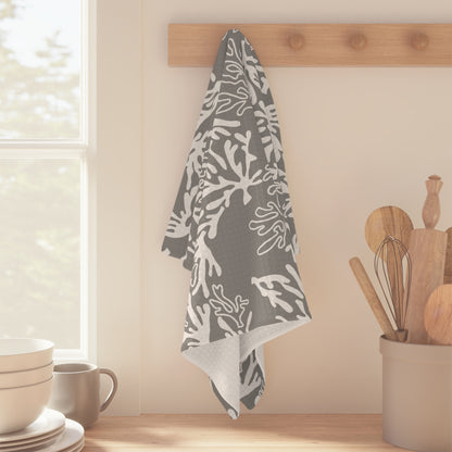 A gray kitchen towel with a white coral pattern hangs from a wooden peg on a kitchen wall, with a window, bowls, and wooden utensils in the background.