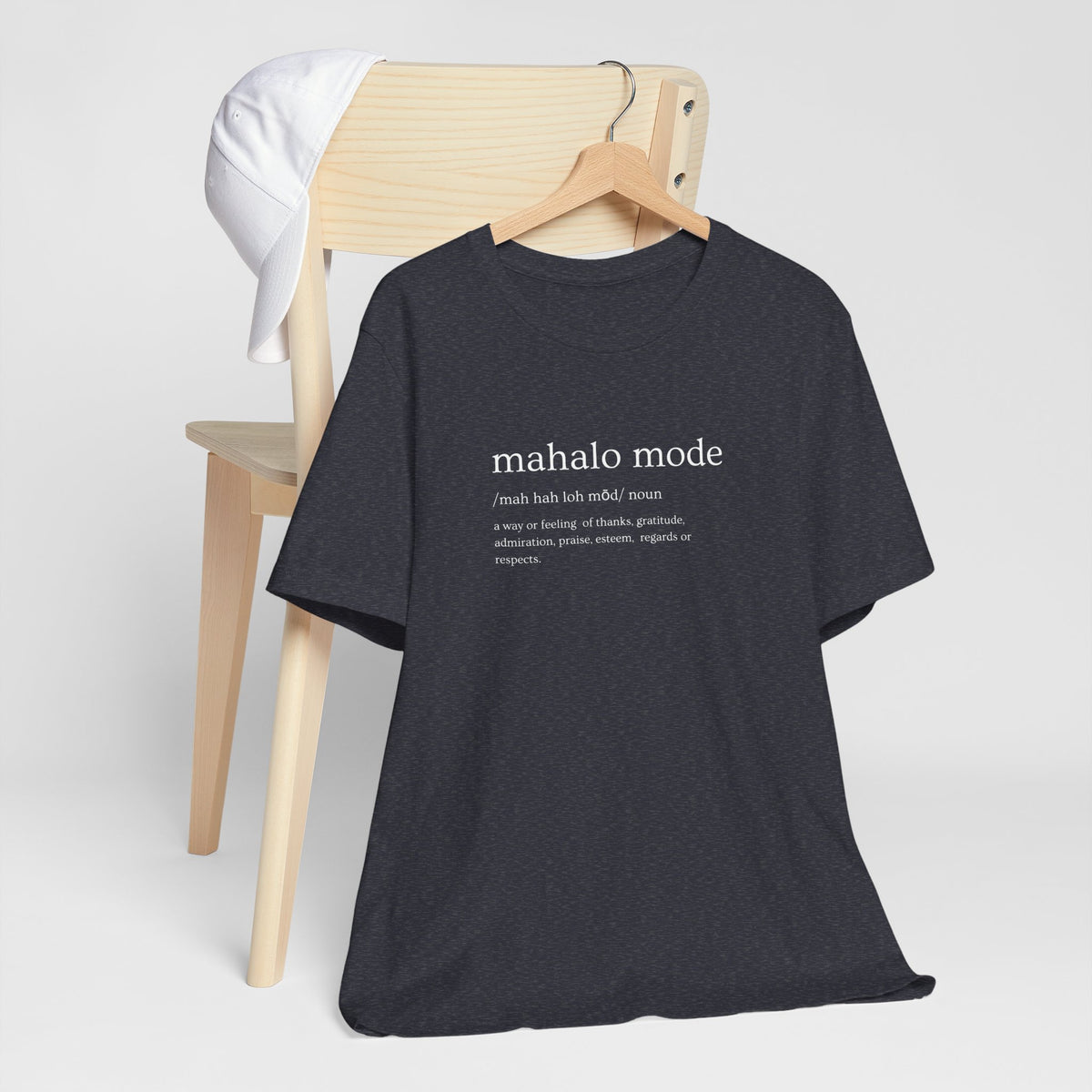 A navy heather t-shirt with a white printed dictionary-style definition of 'mahalo mode' on the chest, hanging from a wooden chair