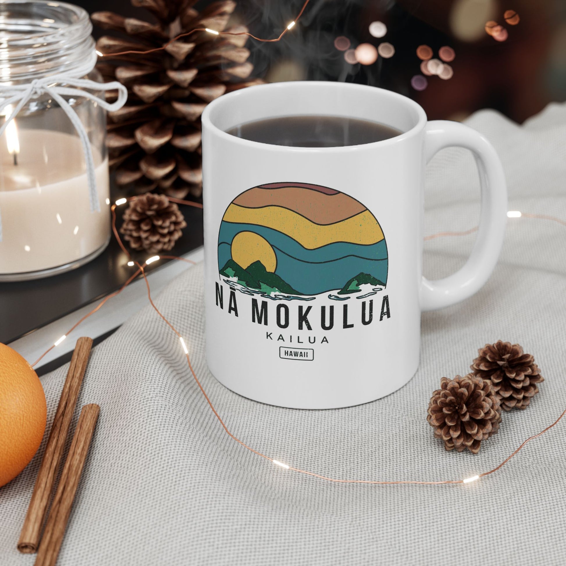 A white ceramic mug with a colorful sunset landscape graphic and the text 'NA MOKULUA KAILUA' on the front, filled with black coffee and styled on a light fabric surface with pinecones, a candle, cinnamon sticks, and string lights.