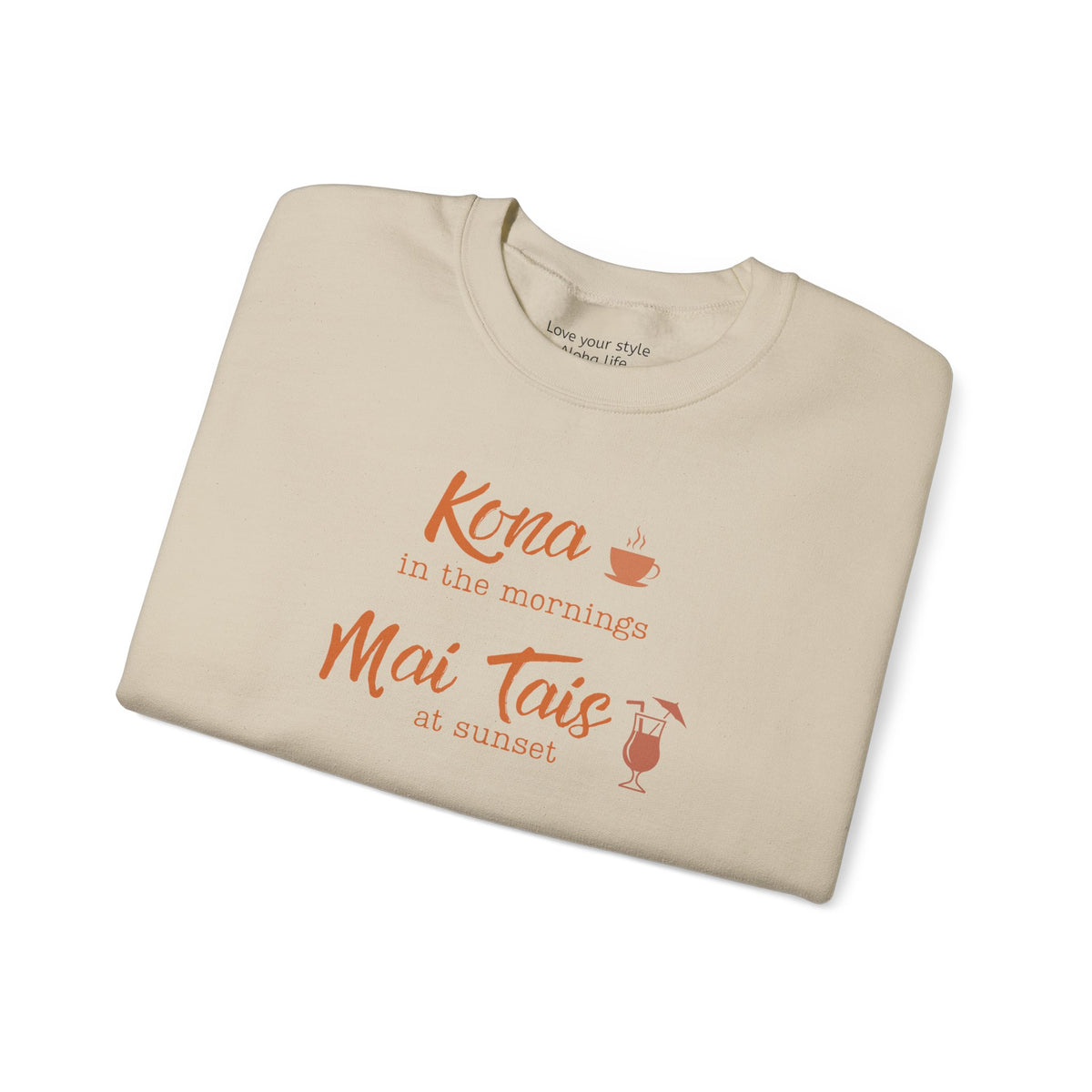 A beige crewneck sweatshirt folded neatly on a white background, featuring orange text that reads 'Kona in the mornings Mai Tais at sunset' with small coffee cup and cocktail glass illustrations.