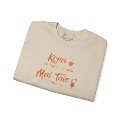 A beige crewneck sweatshirt folded neatly on a white background, featuring orange text that reads 'Kona in the mornings Mai Tais at sunset' with small coffee cup and cocktail glass illustrations.