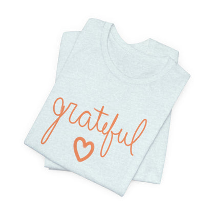 A folded light blue t-shirt with the word 'grateful' written in orange cursive script and a matching orange heart outline on the front, set against a plain white background.