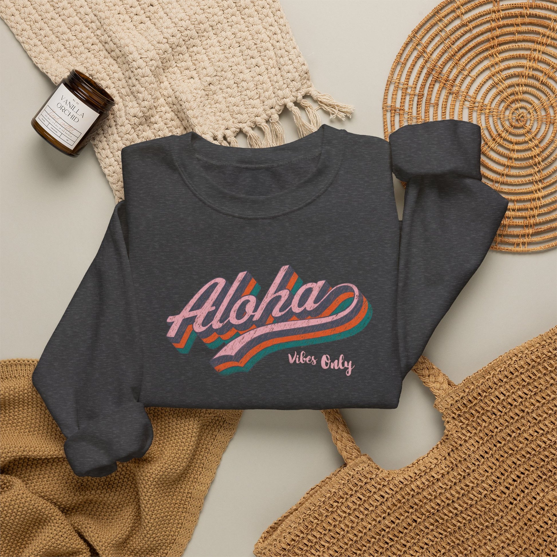 A dark gray crewneck sweatshirt with a retro-style 'Aloha Vibes Only' graphic in pink, orange, and teal, styled in a flat lay with a beige knit blanket, a woven basket, and a candle.