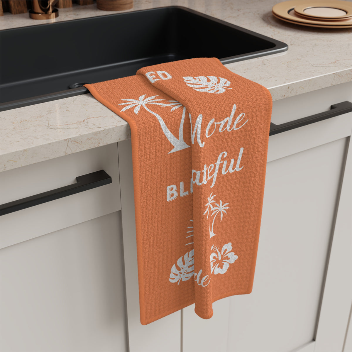 An orange waffle-weave kitchen towel with white tropical prints and text hangs over the edge of a black sink in a modern kitchen setting.