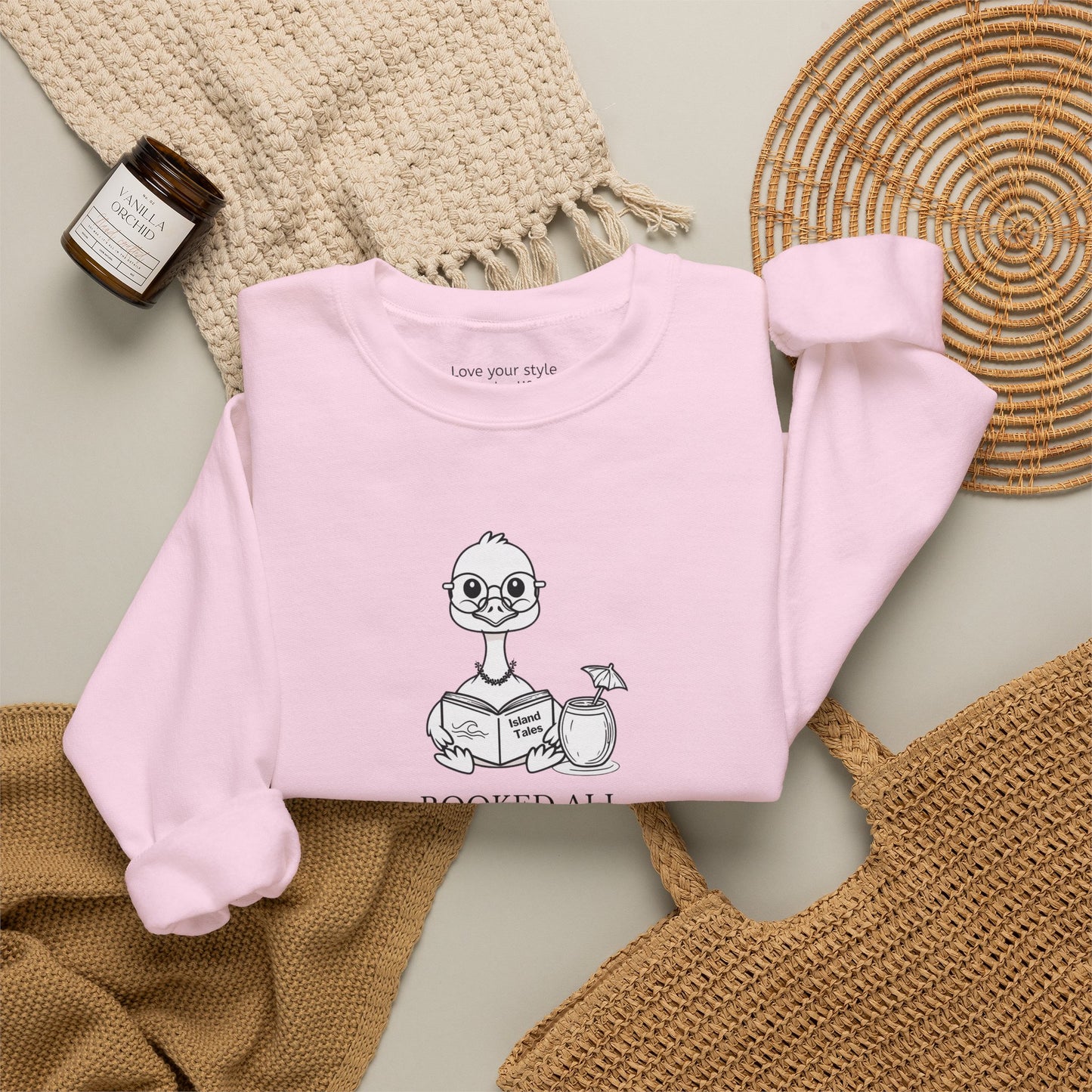 A light pink sweatshirt laid flat on a neutral surface, featuring a cartoon nene goose reading a book and the text 'booked all weekend' on the front. The sweatshirt is styled with a beige knit blanket, a woven straw mat, a small candle jar, and a tan tote bag.