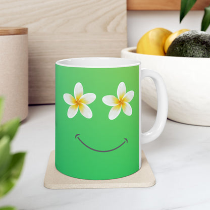 A green ceramic mug with a white handle and interior, decorated with two white plumeria flowers for eyes and a curved smile, sitting on a light wood coaster in a kitchen setting.