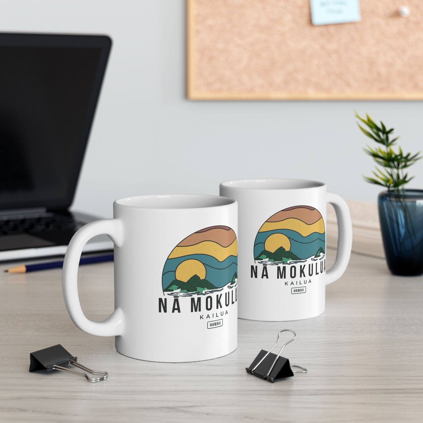 Two white ceramic mugs with a colorful 'Na Mokulua kailua hawaii' sunset graphic, displayed on a light wood desk in a blurred office setting.