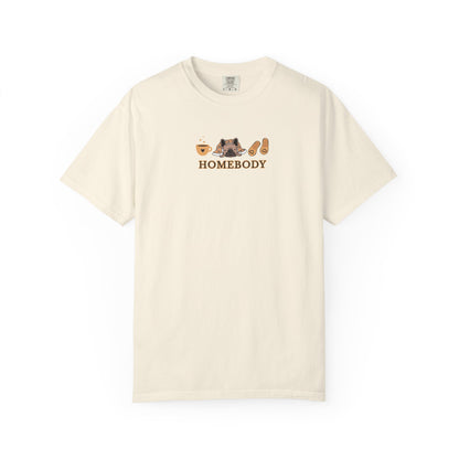An off-white short-sleeve crewneck t-shirt with a graphic on the chest depicting a dog under a blanket, a coffee mug, a pair of house slippers, and the word 'HOMEBODY' in brown text.