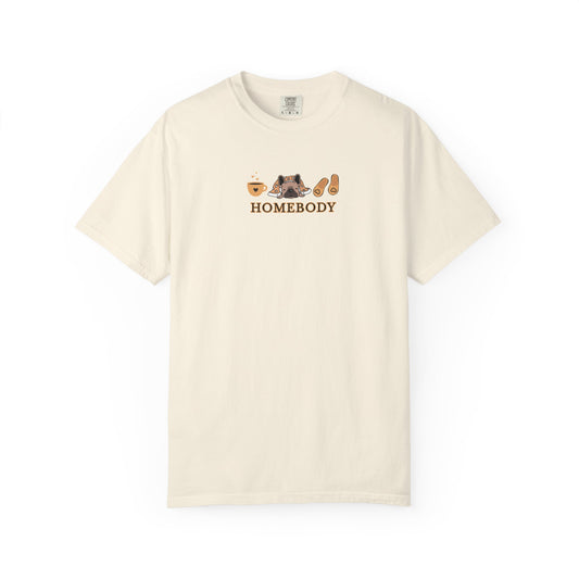 An off-white short-sleeve crewneck t-shirt with a graphic on the chest depicting a dog under a blanket, a coffee mug, a pair of house slippers, and the word 'HOMEBODY' in brown text.