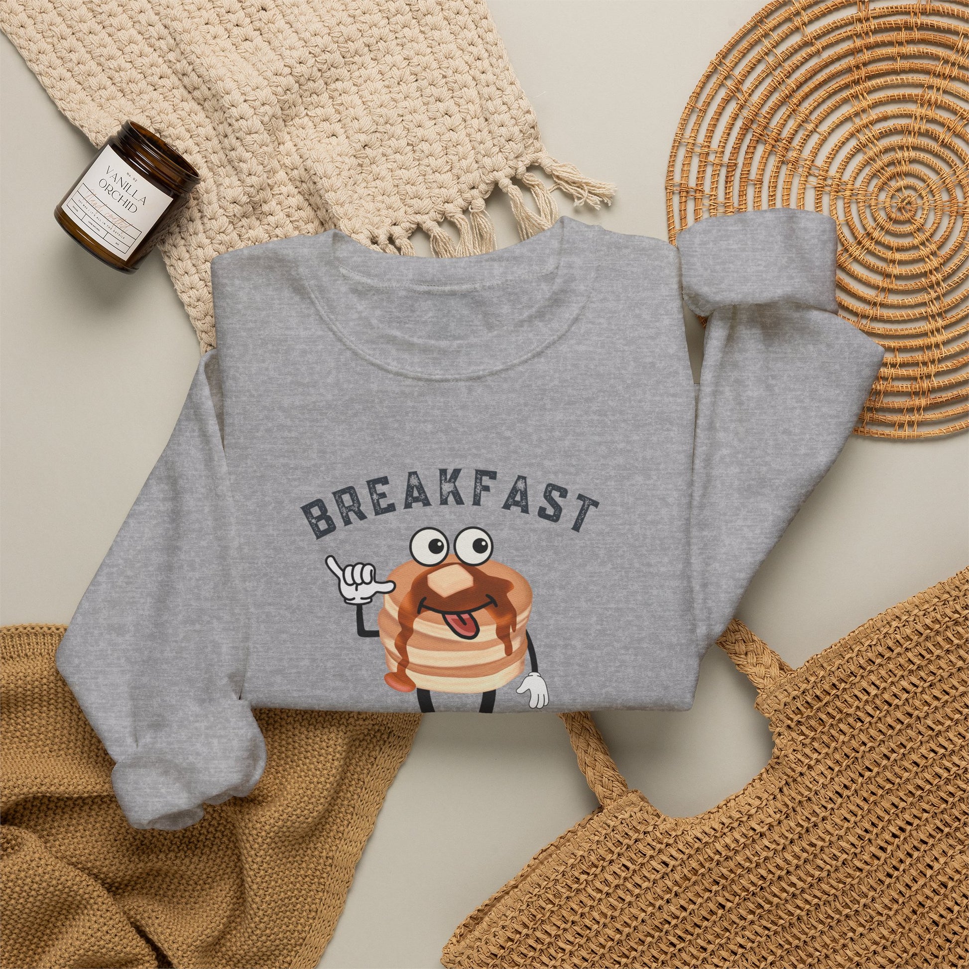 A heather gray sweatshirt laid flat on a light beige surface, featuring a cartoon pancake graphic with the word 'BREAKFAST' above it. The sweatshirt is styled with a cream knit blanket, a small amber candle jar, and woven natural-fiber accessories.