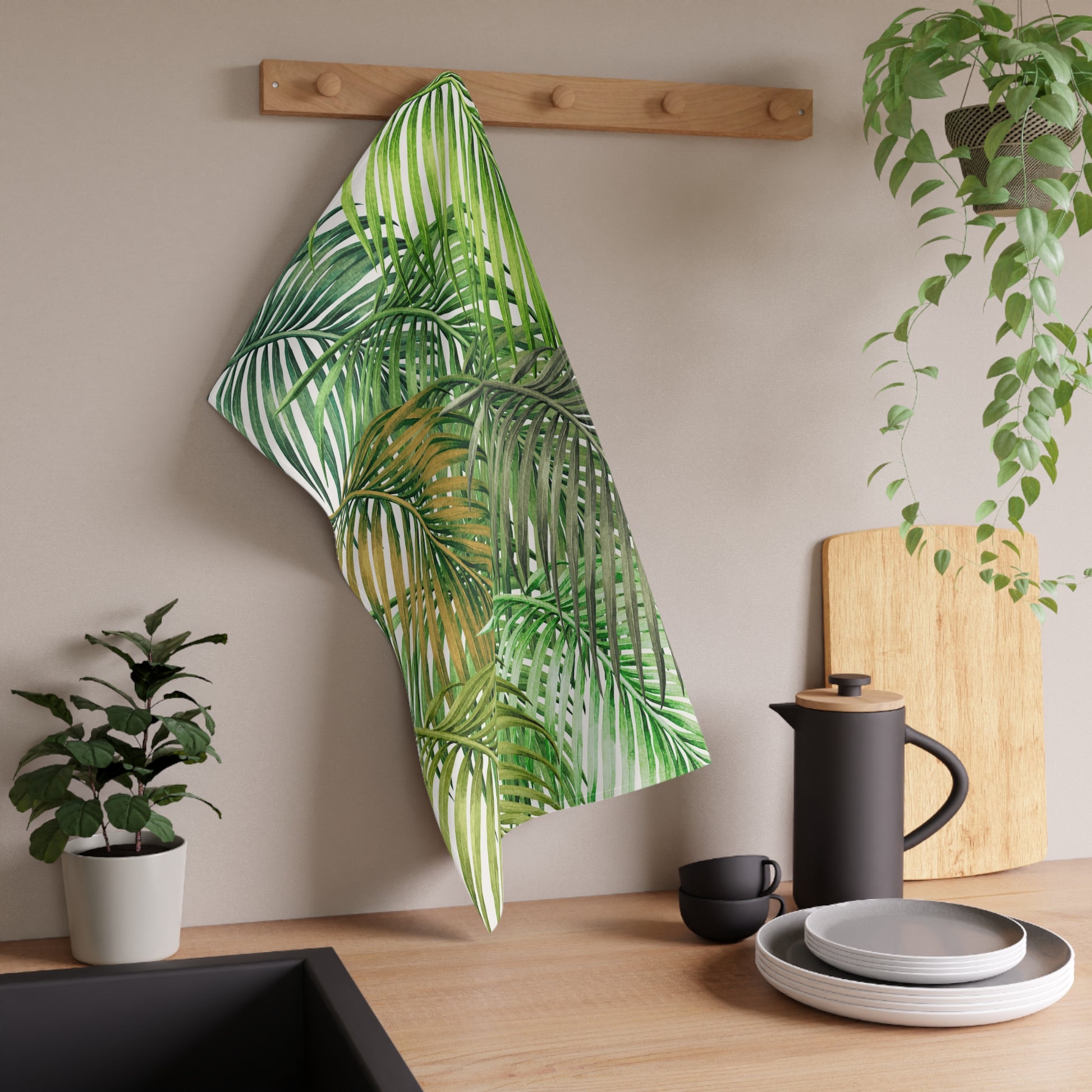 A kitchen towel with a green tropical palm leaf pattern hanging from a wooden peg on a wall in a modern kitchen setting.