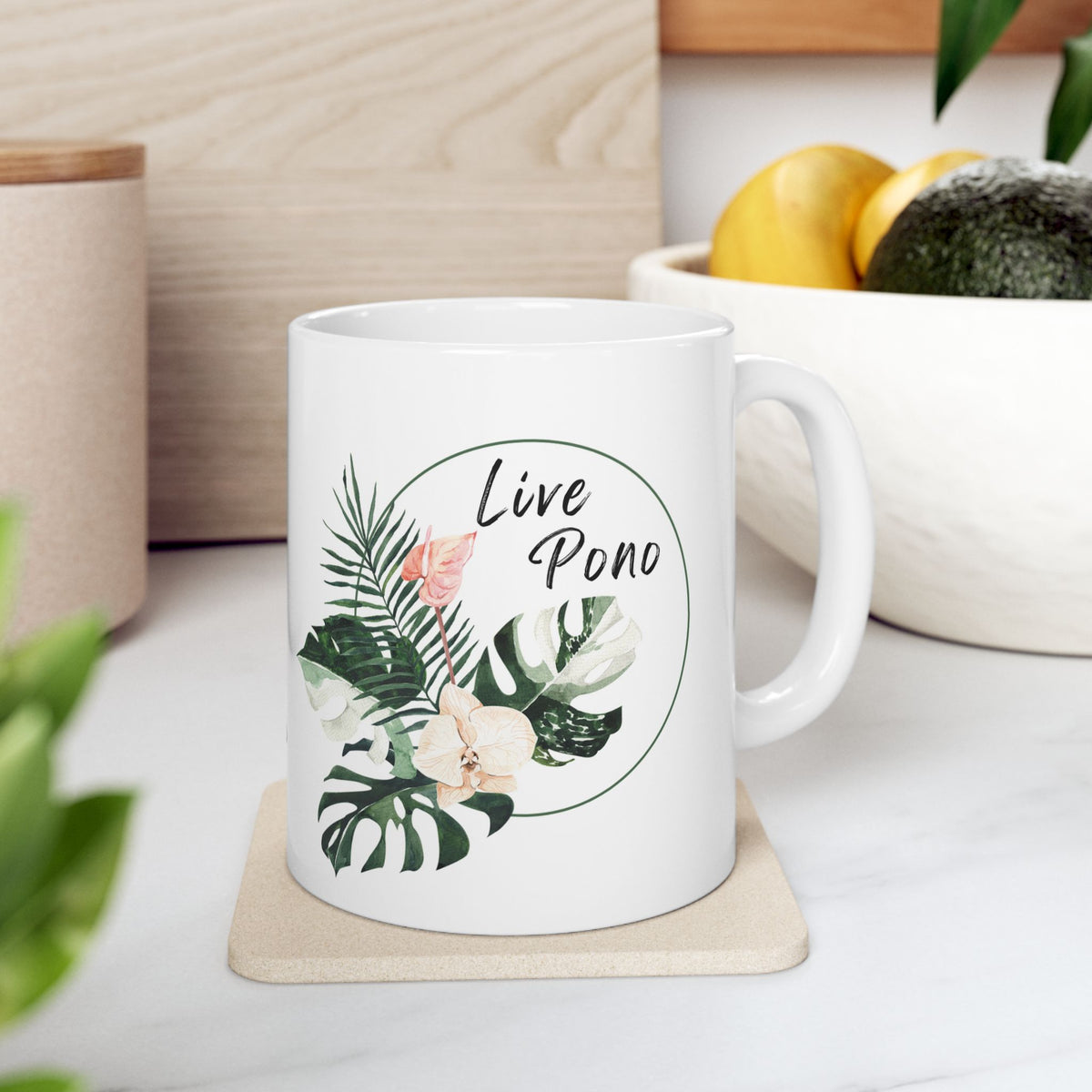 A white ceramic mug with a tropical leaf and flower illustration and the words 'Live Pono' printed on the side, sitting on a beige coaster on a kitchen counter.