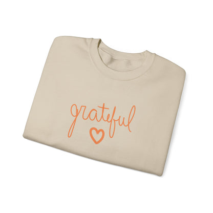 A beige crewneck sweatshirt folded neatly, featuring the word 'grateful' in orange cursive script and a matching orange heart outline on the front, set against a plain white background.