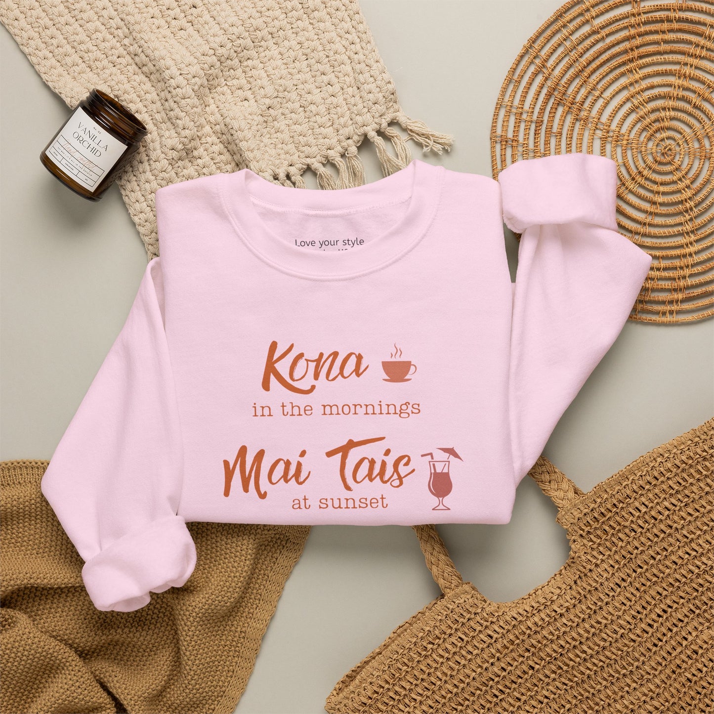 A light pink crewneck sweatshirt folded neatly on natural accessoires featuring orange text that reads 'Kona in the mornings Mai Tais at sunset' with small coffee cup and cocktail glass illustrations.