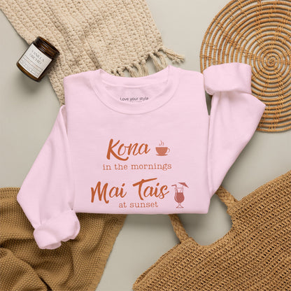 A light pink crewneck sweatshirt folded neatly on natural accessoires featuring orange text that reads 'Kona in the mornings Mai Tais at sunset' with small coffee cup and cocktail glass illustrations.