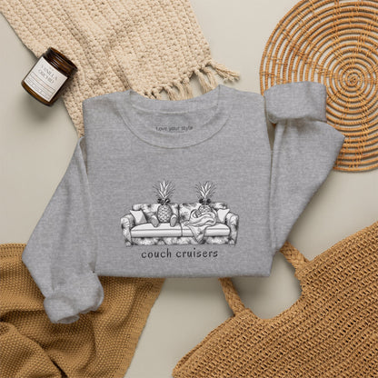 A gray-colored crewneck sweatshirt with a black and white graphic of a couch with pineapples and the text 'couch cruisers', styled in a flat lay with a woven blanket, a candle, and a wicker tray.