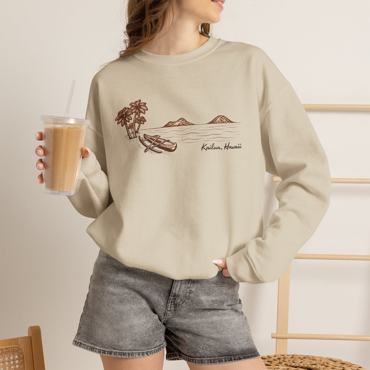 Woman standing wearing Tonal hand-drawn Kailua canoe and Mokulua Islands unisex sweatshirt in warm neutral shades holding an iced coffee.