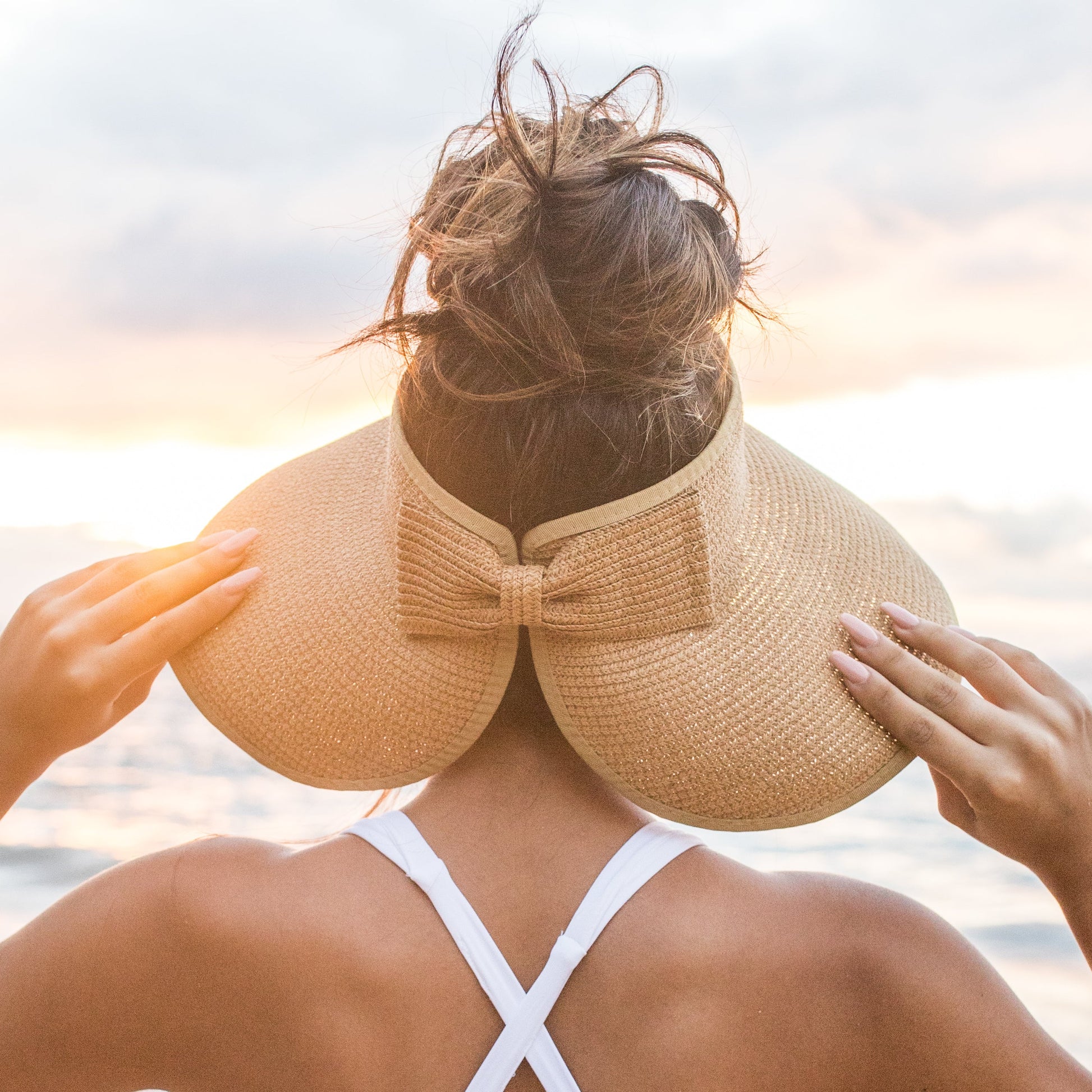 Person wearing a large straw hat with a bow, facing away from the camera with a sunset or sunrise in the background.