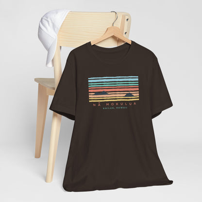 A hanging black t-shirt with a colorful retro sunset graphic and the text 'NÁ MOKULUA KAILUA, HAWAII' on the front, hanging from a wood chair.