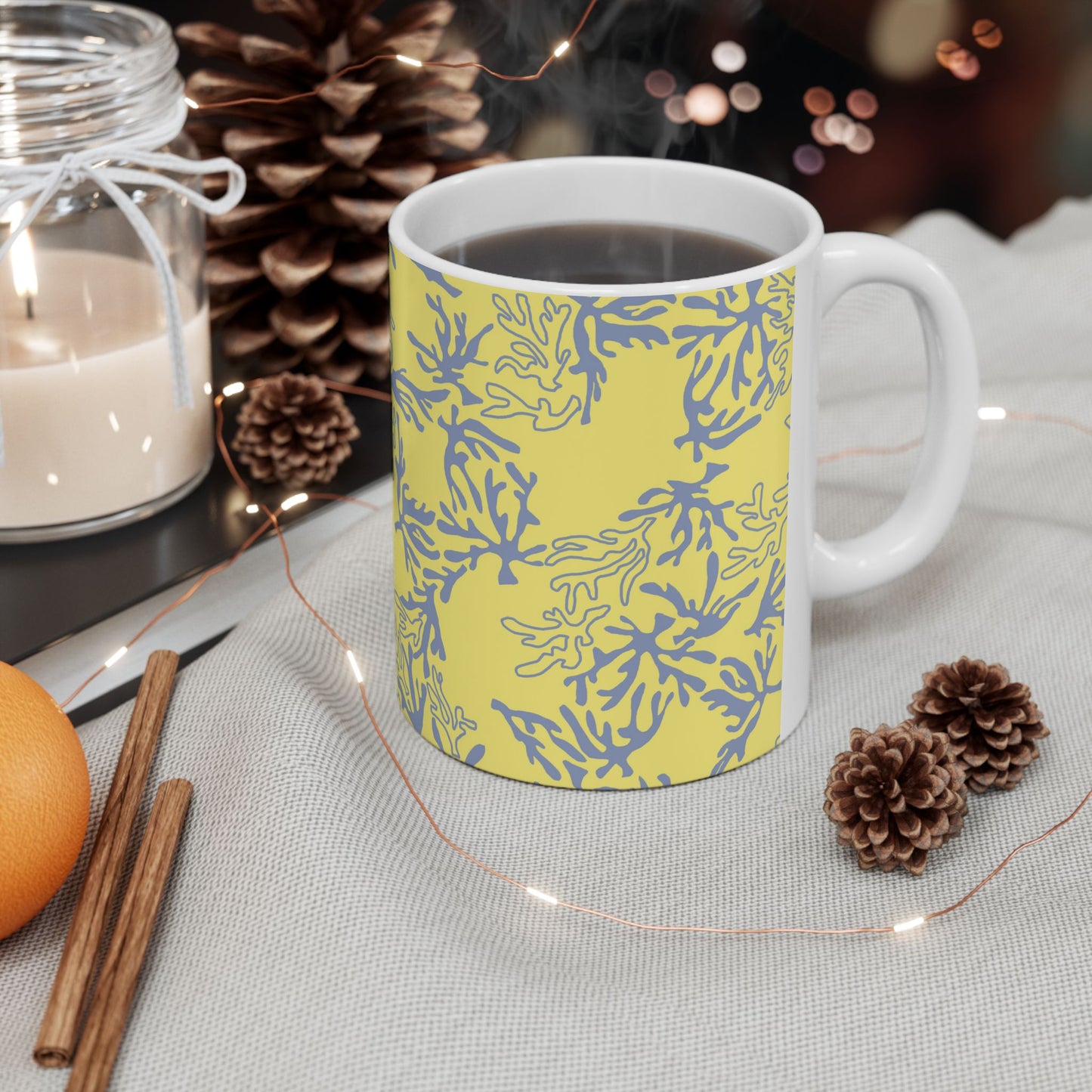 A white ceramic mug filled with a dark beverage, with a yellow grey/blue coral pattern. The mug is styled on a light gray fabric surface with a candle, pinecones, cinnamon sticks, and fairy lights in the background.