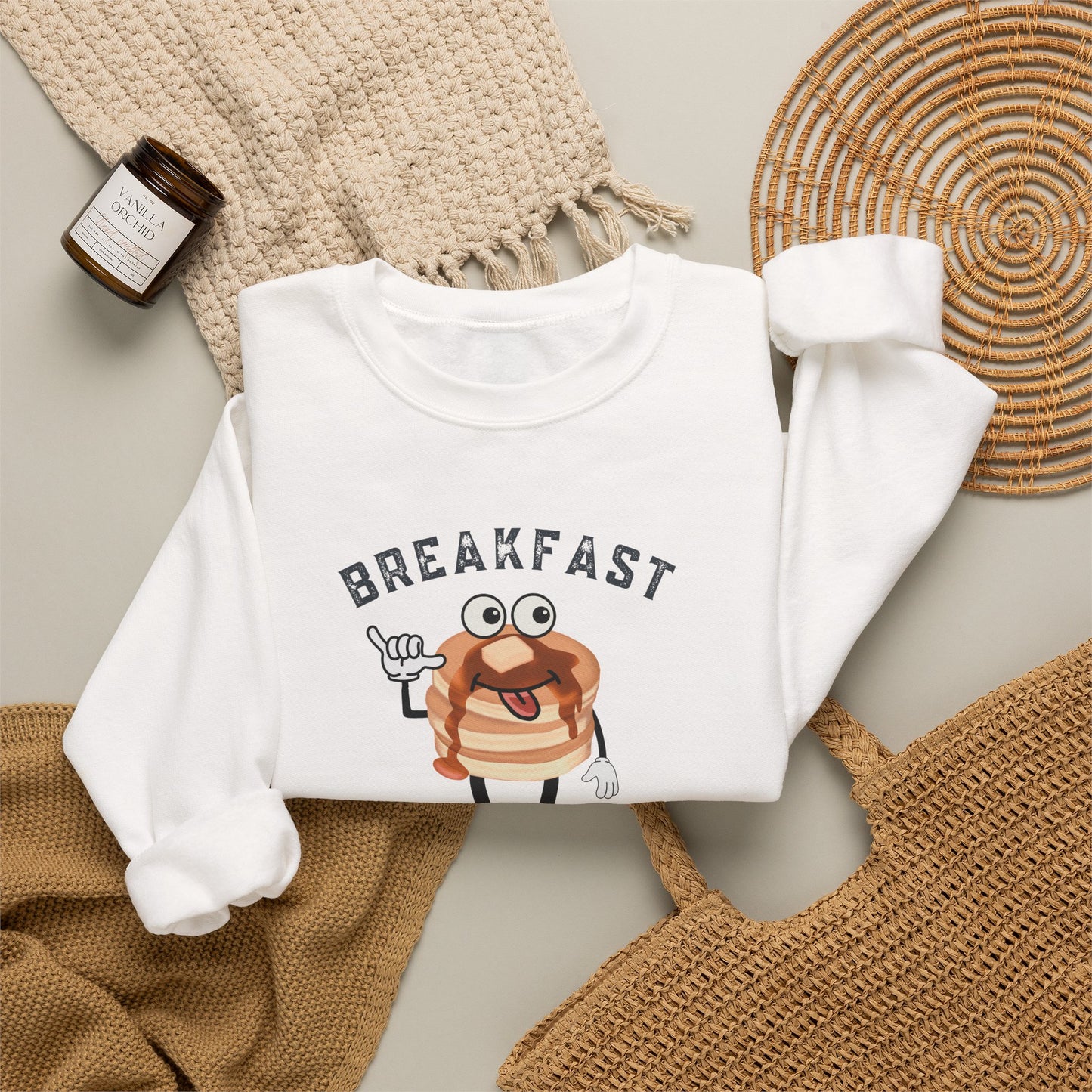 A folded white long-sleeve sweatshirt with a cartoon pancake graphic and the word 'BREAKFAST' printed on the front, styled in a flat lay with a beige knit blanket, woven mat, and small candle.