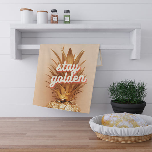 A kitchen towel featuring a golden pineapple graphic and the words 'stay golden' in pink script, hanging on a white wall-mounted rack in a bright kitchen setting.