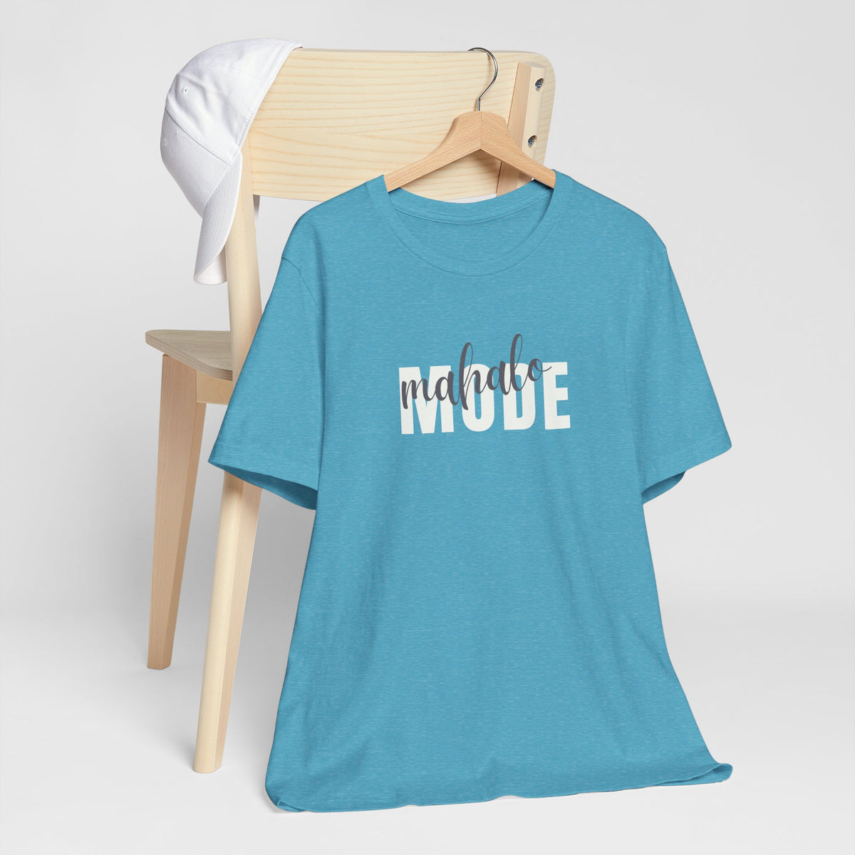 An aqua heather short-sleeve t-shirt with a 'mahalo mode' graphic on the chest, hanging on a wooden hanger from a light wood chair with a white cap hanging on the chair, set against a plain light gray background.