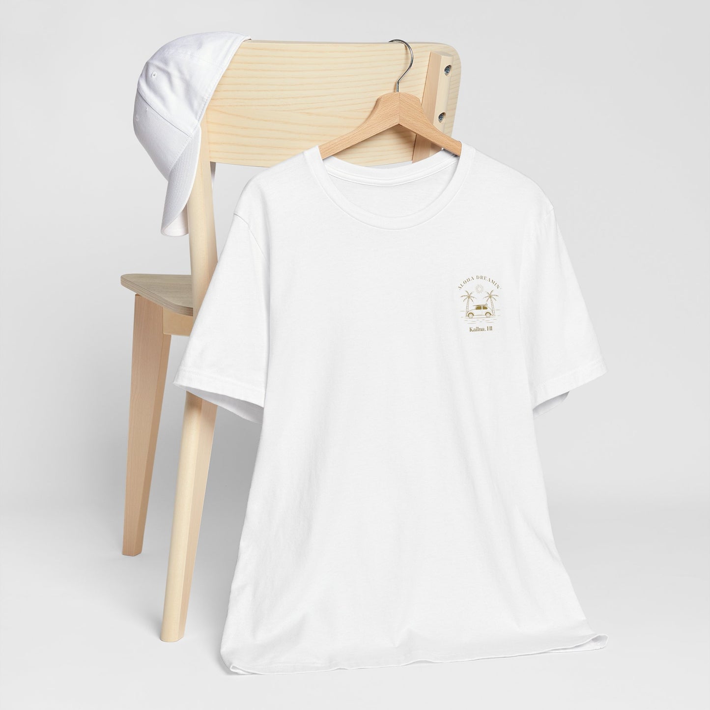 A white crewneck t-shirt with a small gold 'Aloha Dreamin Kailua Hawaii' design on the left chest, displayed on a wooden hanger draped over a light wood chair with a white shirt hanging off the backrest, against a neutral light gray background.