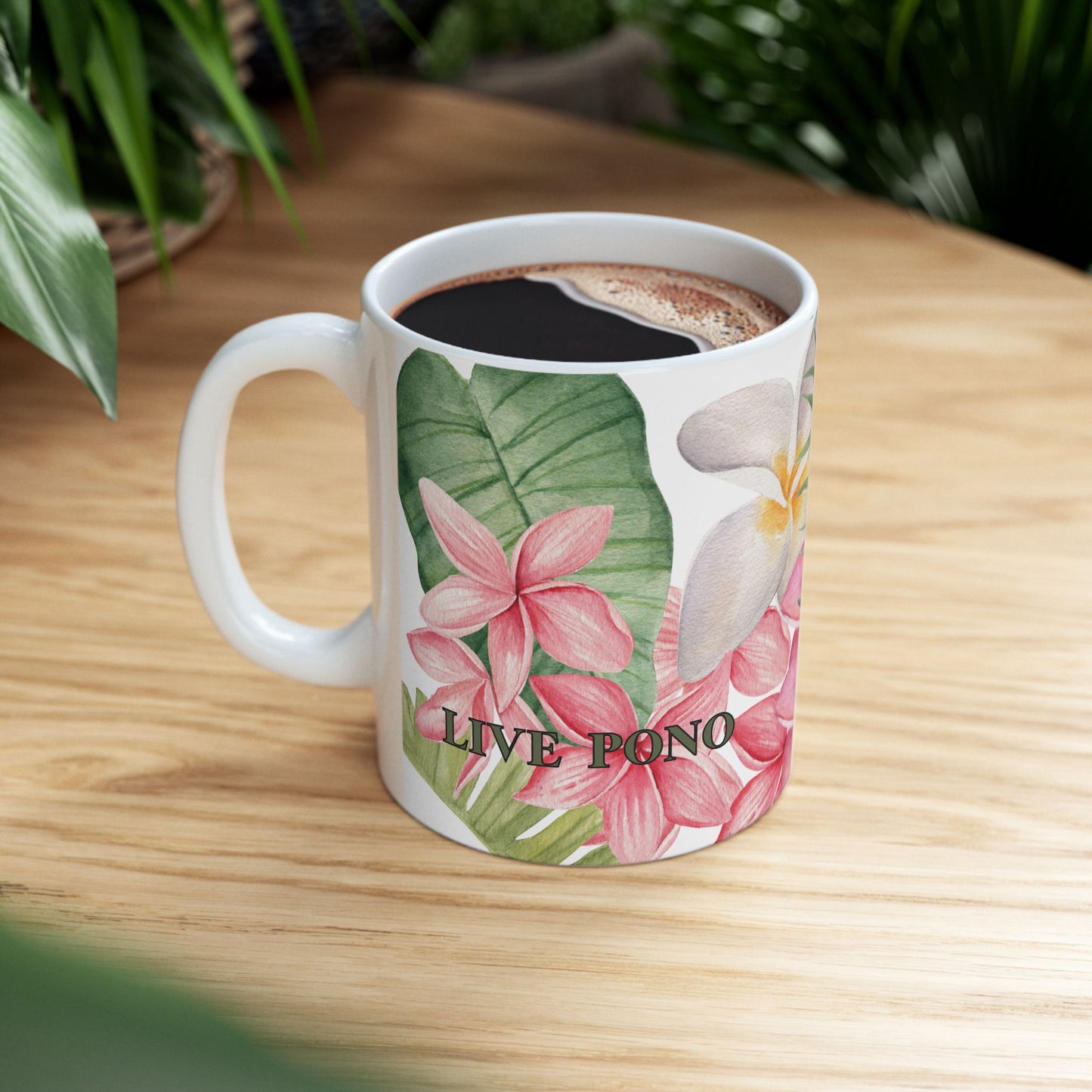 A white ceramic mug with a tropical floral print featuring pink plumeria flowers, green leaves, and the text 'LIVE PONO', filled with a dark beverage and placed on a light wooden table with blurred green plants in the background.