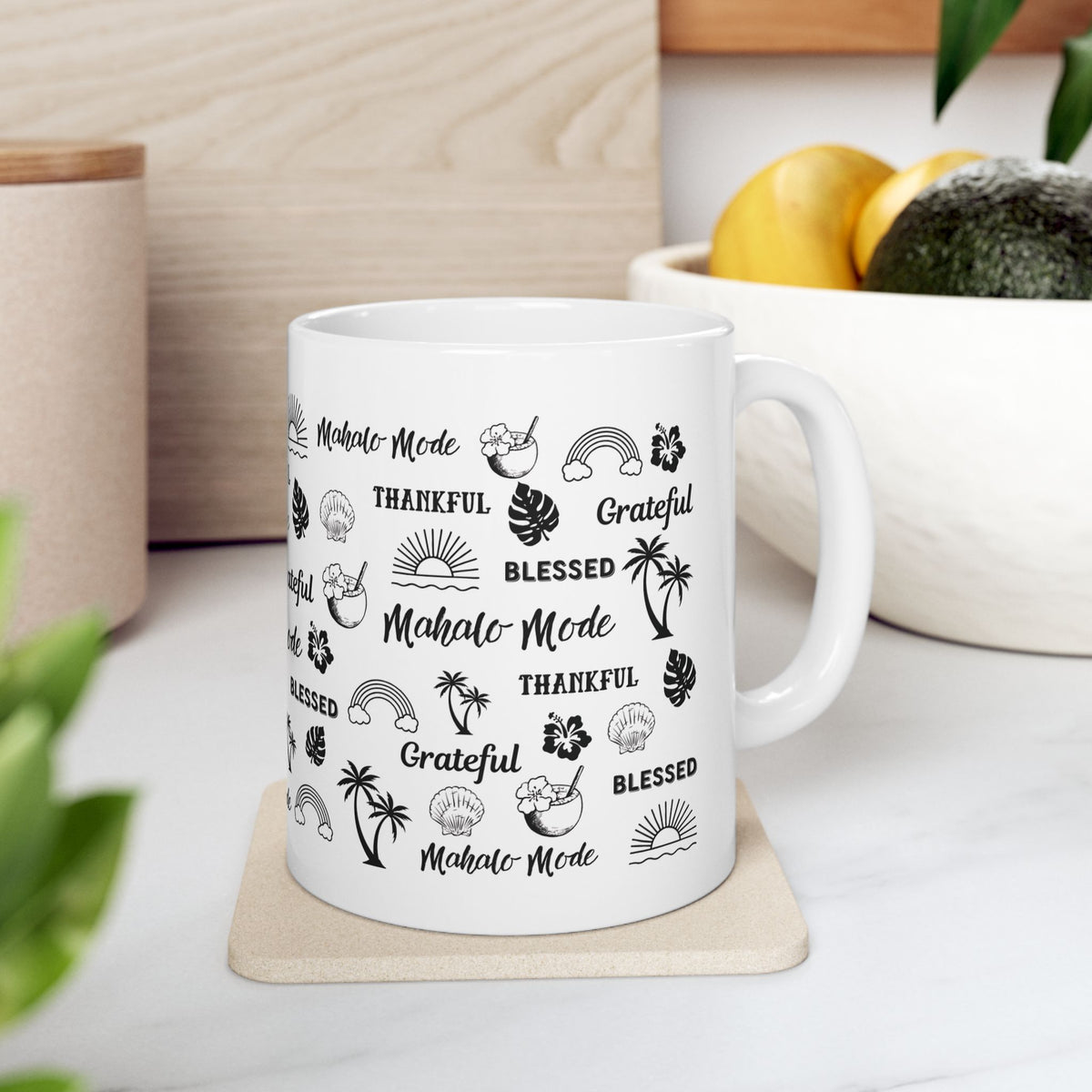 A white ceramic mug with a black tropical-themed print featuring the words 'Mahalo Mode', 'Grateful', and 'Blessed' alongside palm trees, monstera leaves, hibiscus flowers, and sun motifs, shown on a coaster, placed on a surface with a plant and container in the background.
