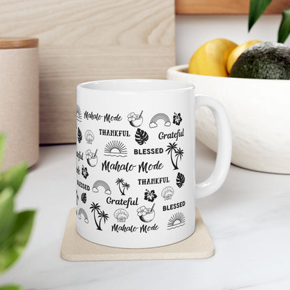 A white ceramic mug with a black tropical-themed print featuring the words 'Mahalo Mode', 'Grateful', and 'Blessed' alongside palm trees, monstera leaves, hibiscus flowers, and sun motifs, shown on a coaster, placed on a surface with a plant and container in the background.