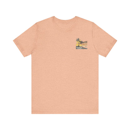 A heather peach short-sleeve t-shirt with a vintage-style graphic on the front left chest depicting two palm trees against a yellow and orange sunset, with the text 'ná makulua' and 'Kailua, Hawaii!' printed below.