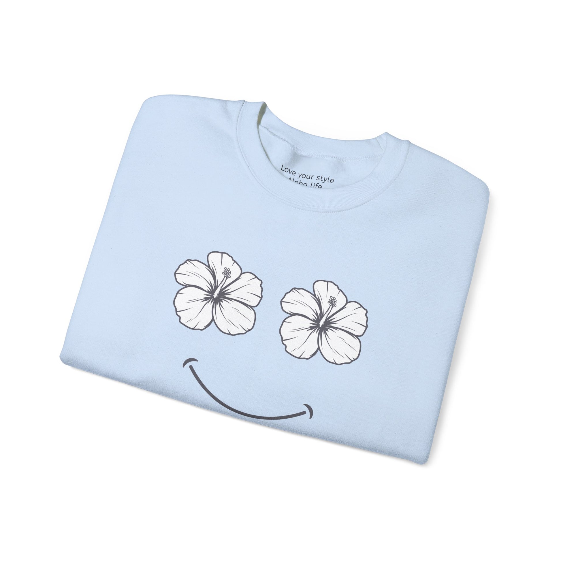 A folded light blue t-shirt with a graphic print of two white hibiscus flowers forming eyes and a black smiley mouth, shown on a plain white background.