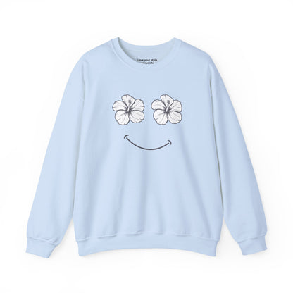 A light blue crewneck sweatshirt featuring a graphic print of two white hibiscus flowers forming eyes and a black smiley mouth on the front.