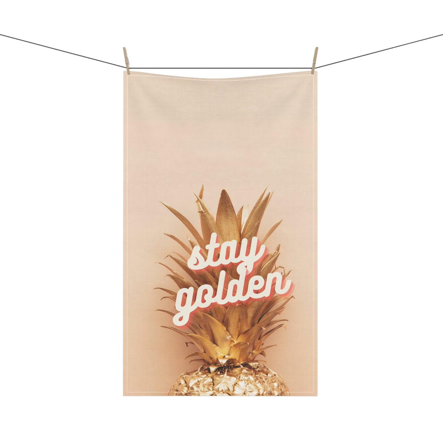 A beige fabric hanging tea towel clipped to a clothesline, featuring a graphic of a golden pineapple with the words 'stay golden' in white script outlined in pink.