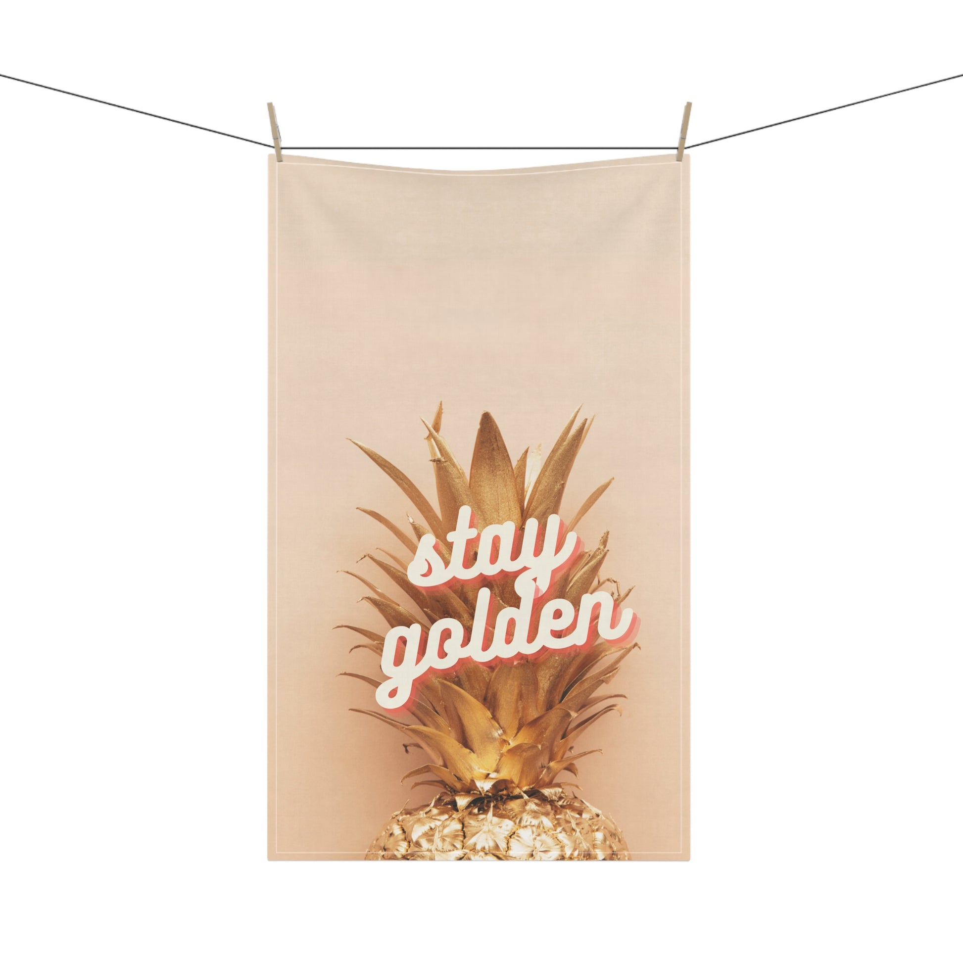 A beige fabric hanging tea towel clipped to a clothesline, featuring a graphic of a golden pineapple with the words 'stay golden' in white script outlined in pink.