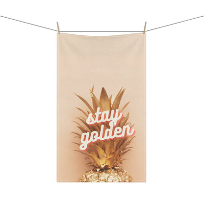 A beige fabric hanging tea towel clipped to a clothesline, featuring a graphic of a golden pineapple with the words 'stay golden' in white script outlined in pink.