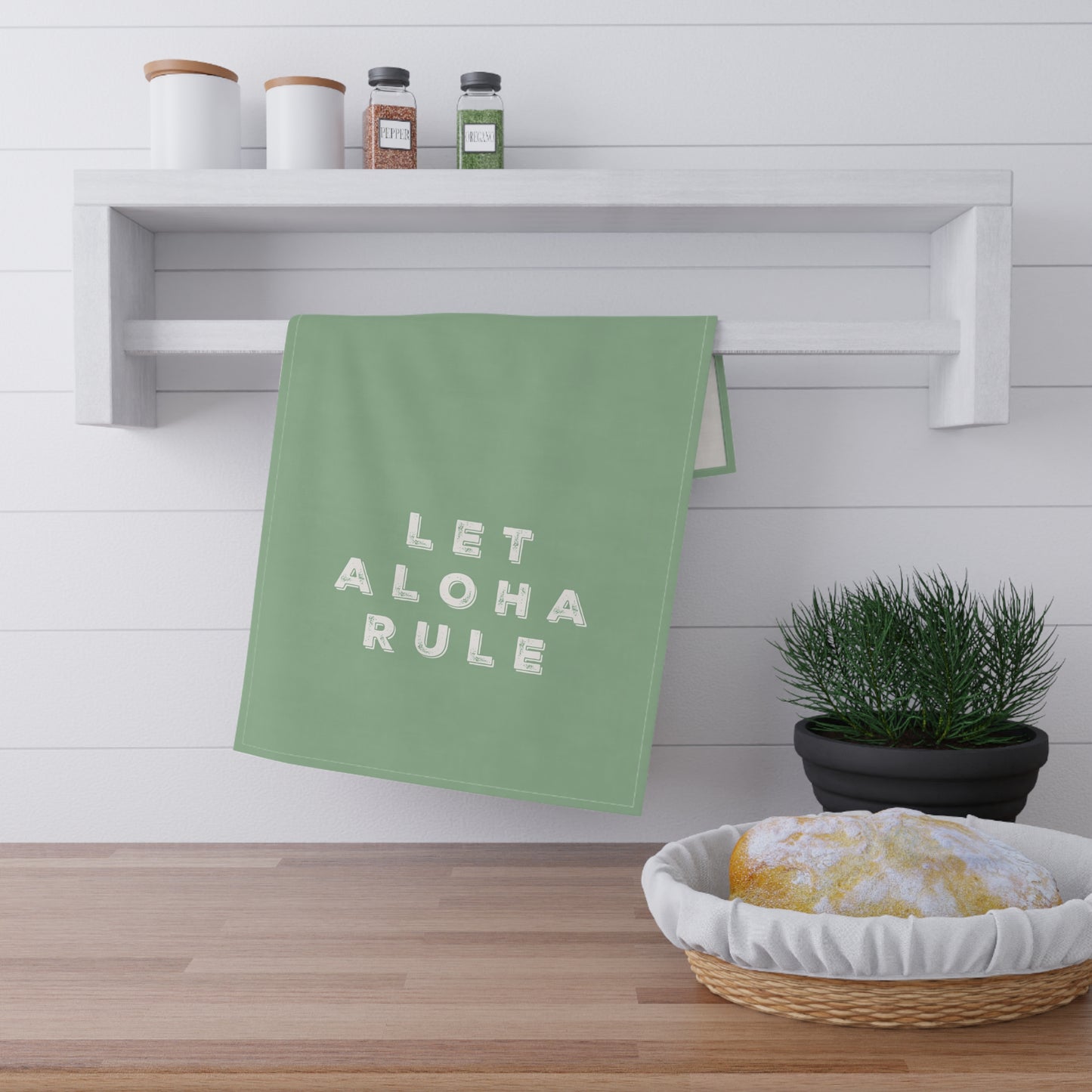 A sage green kitchen towel with the words 'LET ALOHA RULE' in white block letters hangs from a white wall-mounted rack. The scene is styled like a kitchen with a wooden floor, spice jars on a shelf, a small potted plant, and a basket with a loaf of bread.