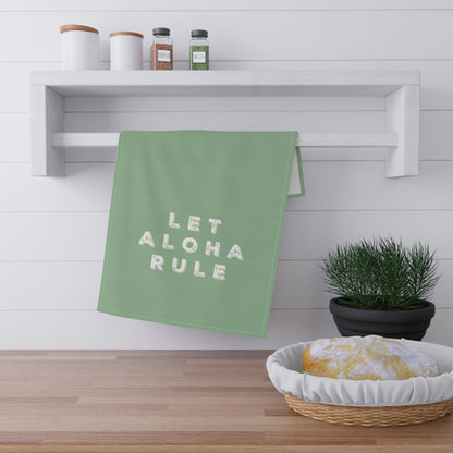 A sage green kitchen towel with the words 'LET ALOHA RULE' in white block letters hangs from a white wall-mounted rack. The scene is styled like a kitchen with a wooden floor, spice jars on a shelf, a small potted plant, and a basket with a loaf of bread.